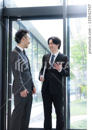 A young businessman and his boss converse by the window in the office surrounded by fresh greenery, discussing business negotiations with clients and their growth. A young businessman and his boss converse by the window in the office surrounded by fresh greenery, discussing business negotiations with clients and their growth. 139542507