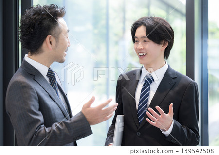 A young businessman and his boss converse by the window in the office surrounded by fresh greenery, discussing business negotiations with clients and their growth. A young businessman and his boss converse by the window in the office surrounded by fresh greenery, discussing business negotiations with clients and their growth. 139542508