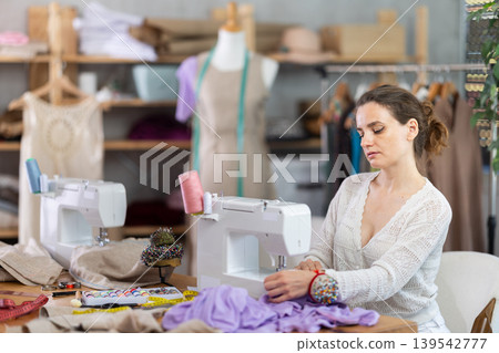 Working in sewing workshop - female dressmaker working at sewing machine making stitches on fabric Working in sewing workshop - female dressmaker working at sewing machine making stitches on fabric 139542777