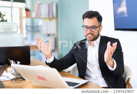 young man in glasses sits at a table and looks at a laptop screen 139542907