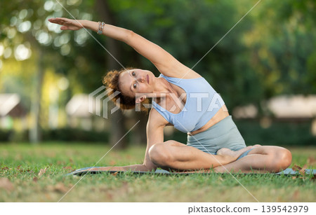 Woman practicing hatha yoga in garden, stretching in an asymmetrical seated asana Parivritta Janu Sirsasana 139542979