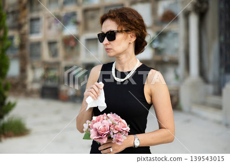 Sad woman in black dress with bouquet of flowers and handkerchief in hands, stands in cemetery 139543015