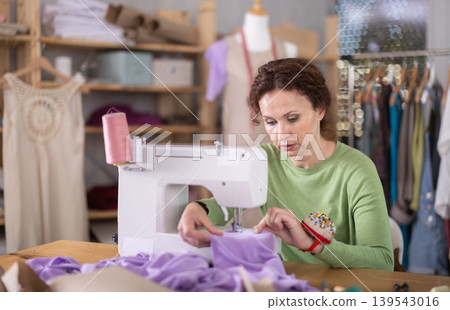 Working in sewing workshop - female dressmaker working at sewing machine making stitches on fabric 139543016