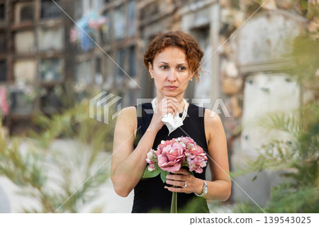 Sad woman in black dress with bouquet of flowers and handkerchief in hands, stands in cemetery 139543025