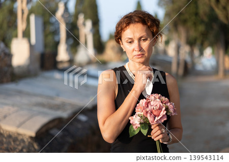 Sad woman in black dress with bouquet of flowers and handkerchief in hands, stands in cemetery Sad woman in black dress with bouquet of flowers and handkerchief in hands, stands in cemetery 139543114