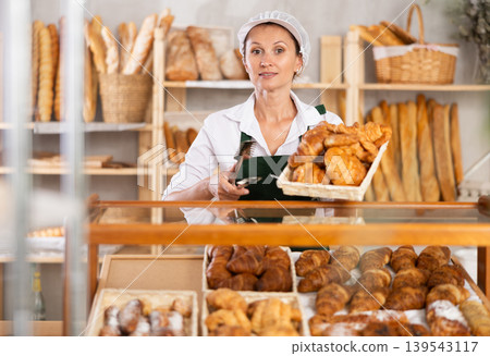 Woman employee puts croissants in window, arranges display of goods at bakery. Woman employee puts croissants in window, arranges display of goods at bakery. 139543117