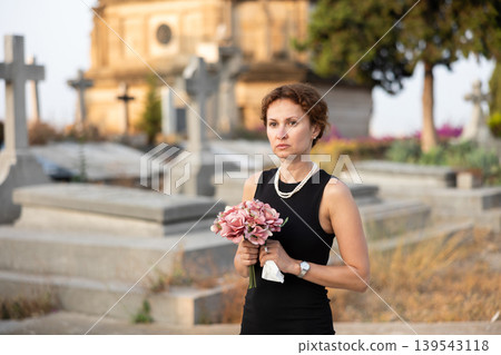 Sad woman in black dress with bouquet of flowers and handkerchief in hands, stands in cemetery 139543118