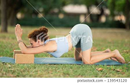 Woman doing yoga in park - sits using wooden blocks practices different yoga poses Woman doing yoga in park - sits using wooden blocks practices different yoga poses 139543120