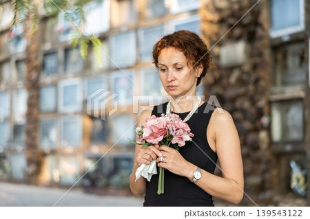 Sad woman in black dress with bouquet of flowers and handkerchief in hands, stands in cemetery 139543122