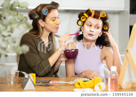 Relaxed female friends applying facial mask to each other at home Relaxed female friends applying facial mask to each other at home 139543139