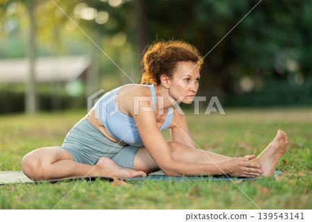 Woman practicing hatha yoga in garden, stretching in an asymmetrical seated asana Parivritta Janu Sirsasana Woman practicing hatha yoga in garden, stretching in an asymmetrical seated asana Parivritta Janu Sirsasana 139543141