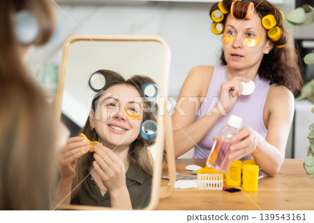 Woman in colorful curlers applying under eye patches by mirror with bestie Woman in colorful curlers applying under eye patches by mirror with bestie 139543161