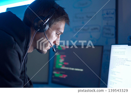Software developer analyzing code on computer monitors while wearing headset in dark workspace Software developer analyzing code on computer monitors while wearing headset in dark workspace 139543329