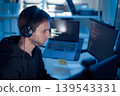 Software developer analyzing code on computer monitors while wearing headset in dark workspace 139543331