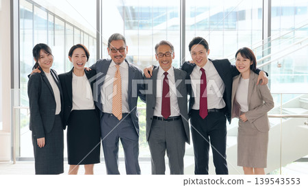 A group of business people of various ages, men and women, with their arms around each other's shoulders, in the lobby. A group of business people of various ages, men and women, with their arms around each other's shoulders, in the lobby. 139543553