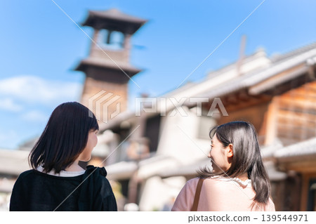 Two young women walking with the Kawagoe Toki no Kane (Time Bell) in the background. Girls' trip, strolling around town, sightseeing in Saitama. Two young women walking with the Kawagoe Toki no Kane (Time Bell) in the background. Girls' trip, strolling around town, sightseeing in Saitama. 139544971