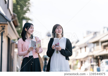 A picture of two women on a girls' trip, walking through the traditional storehouse district of Kawagoe, Saitama, holding tourist brochures. A picture of two women on a girls' trip, walking through the traditional storehouse district of Kawagoe, Saitama, holding tourist brochures. 139544975