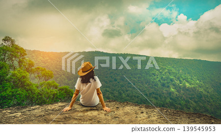 Young Asian woman traveler wearing hat sitting on mountain cliff, relaxing and watching forest view during solo vacation, outdoor adventure. Freedom, peaceful lifestyle and nature exploration. Young Asian woman traveler wearing hat sitting on mountain cliff, relaxing and watching forest view during solo vacation, outdoor adventure. Freedom, peaceful lifestyle and nature exploration. 139545953