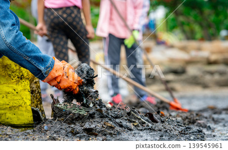Adult and children volunteers collecting garbage on the sea beach. Beach environment pollution. Tidying up rubbish on beach. People wear orange gloves picking garbage up in to yellow bag. 139545961