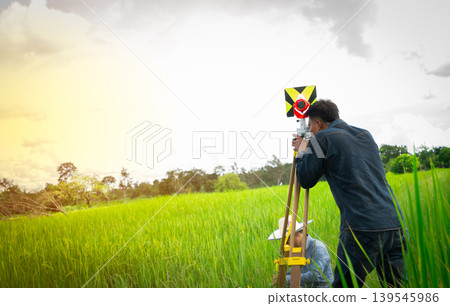 Asian engineer using theodolite to measure rice field land, surveyor working in rural farmland. Land surveying, agricultural planning, infrastructure development and precision engineering fieldwork. Asian engineer using theodolite to measure rice field land, surveyor working in rural farmland. Land surveying, agricultural planning, infrastructure development and precision engineering fieldwork. 139545986