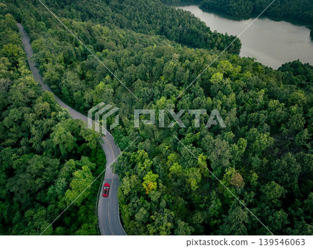 Aerial top view of a red car driving on highway road in green forest. Sustainable transport. Electric vehicle driving on asphalt road through lush green forest. Zero emission car. Green mobility. Aerial top view of a red car driving on highway road in green forest. Sustainable transport. Electric vehicle driving on asphalt road through lush green forest. Zero emission car. Green mobility. 139546063