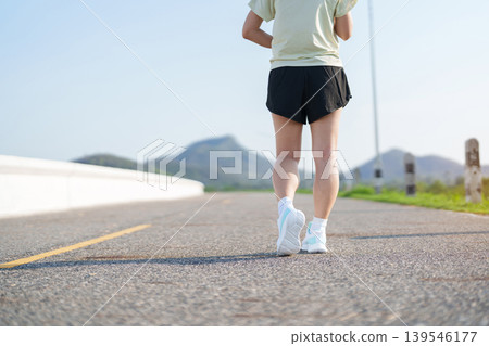Young adult Asian woman jogging and walking in the park at morning. Female athlete in sport shoes running on the road with visible leg muscles. Healthy lifestyle, wellness and workout concept Young adult Asian woman jogging and walking in the park at morning. Female athlete in sport shoes running on the road with visible leg muscles. Healthy lifestyle, wellness and workout concept 139546177