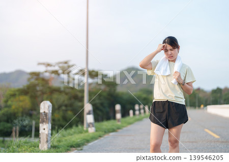 Exhausted Asian woman wiping sweat with a towel after jogging in the park. Tired female runner suffering from summer heat and fatigue after fitness workout. Outdoor exercise and heatstroke concept Exhausted Asian woman wiping sweat with a towel after jogging in the park. Tired female runner suffering from summer heat and fatigue after fitness workout. Outdoor exercise and heatstroke concept 139546205