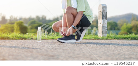 Asian man with towel tying running shoes in the park with a reusable water bottle. male runner getting ready for jogging and fitness exercise. Healthy lifestyle and sustainability concept 139546237