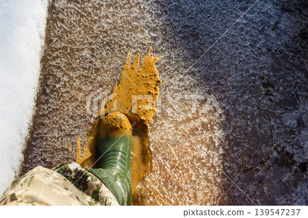 Green rubber boot sploshing through a partially frozen stream, breaking ice and splashing muddy slush. Early spring exploration Green rubber boot sploshing through a partially frozen stream, breaking ice and splashing muddy slush. Early spring exploration 139547237