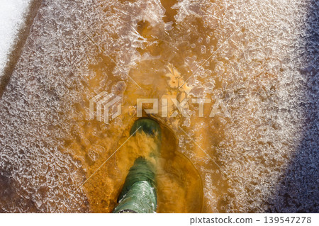 Green rubber boot in frozen creek, stomping through fragile ice and murky water. A March adventure 139547278