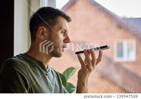 Man using voice assistant on smartphone by window at home Man using voice assistant on smartphone by window at home 139547589