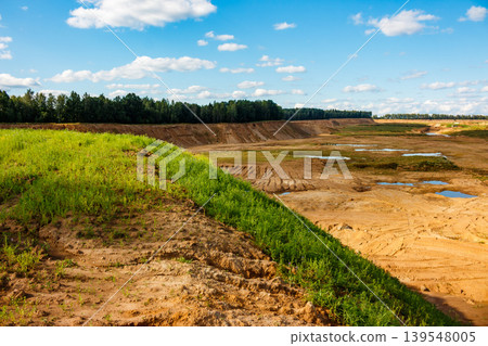 Vibrant green grassy slope overlooks an expansive sand pit, flanked by distant forest under a bright blue sky Vibrant green grassy slope overlooks an expansive sand pit, flanked by distant forest under a bright blue sky 139548005