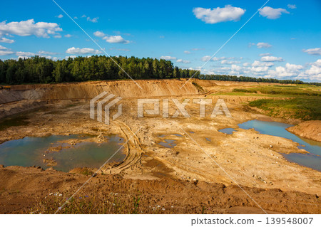 Expansive sand quarry, a large dig site showing vehicle tracks, sandy slopes, and shallow water pools. Massive earthwork under a bright sky Expansive sand quarry, a large dig site showing vehicle tracks, sandy slopes, and shallow water pools. Massive earthwork under a bright sky 139548007