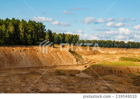 Vast sand quarry with steep, eroded slopes and sandy terrain. Dense forest borders the pit under a bright sky, showcasing an open-cast landscape Vast sand quarry with steep, eroded slopes and sandy terrain. Dense forest borders the pit under a bright sky, showcasing an open-cast landscape 139548010
