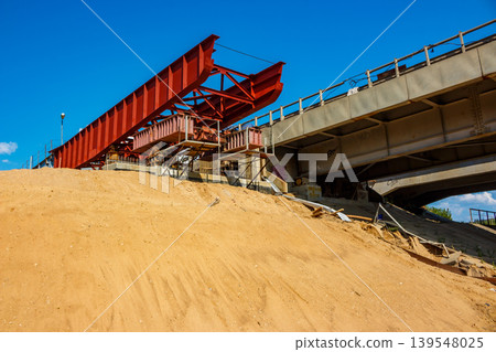 Massive red gantry system under construction, towering over a sandy embankment beneath a clear blue sky. New infrastructure build 139548025