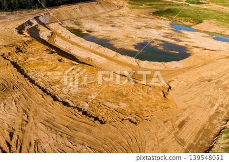 Expansive sand pit, an open-cast mine, showing tiered excavation work. Barren earth, fresh tire marks, and scattered water pools reflect active aggregate extraction operations Expansive sand pit, an open-cast mine, showing tiered excavation work. Barren earth, fresh tire marks, and scattered water pools reflect active aggregate extraction operations 139548051