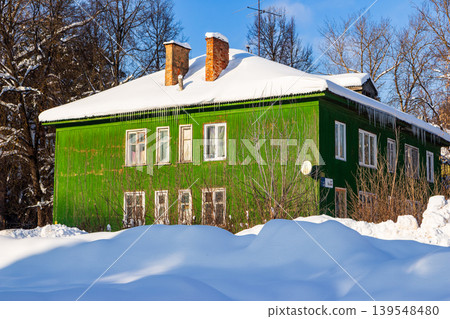 Charming emerald abode blanketed in fresh winter snow, its roof adorned with shimmering icicles. Bright sunny day illuminating the rustic facade against a crisp blue sky 139548480