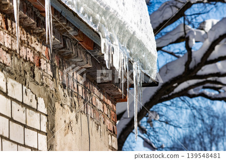 Sparkling icicles dangle from a snow-laden roof against an old brick wall. Winter's icy beauty catches sunlight, creating a frosty, tranquil scene 139548481