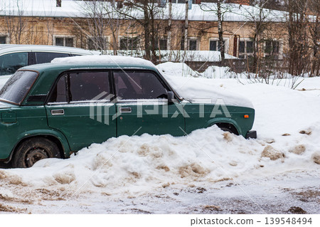 Classic green sedan, caked in fresh snow and dirty slush, hunkers down on a bleak winter street. Urban backdrop adds a touch of vintage charm, Russia Classic green sedan, caked in fresh snow and dirty slush, hunkers down on a bleak winter street. Urban backdrop adds a touch of vintage charm, Russia 139548494