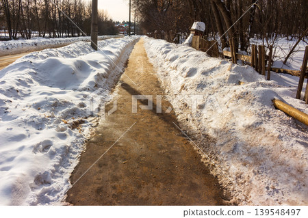 Gritty, damp path cuts through melting winter snowbanks, flanked by bare trees and rustic fence. Bright sun illuminates transitional season walk Gritty, damp path cuts through melting winter snowbanks, flanked by bare trees and rustic fence. Bright sun illuminates transitional season walk 139548497