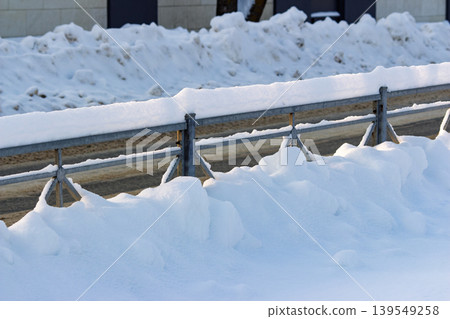 Metal guardrail heavily laden with pristine white snow drifts against a backdrop of winter landscape and cleared roadway Metal guardrail heavily laden with pristine white snow drifts against a backdrop of winter landscape and cleared roadway 139549258