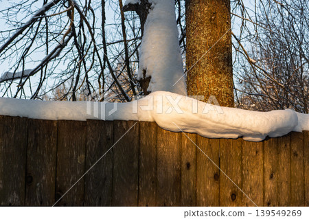 Thick blanket of fresh snow rests atop a weathered wooden fence under warm evening sunlight casting a golden glow on a winter scene 139549269