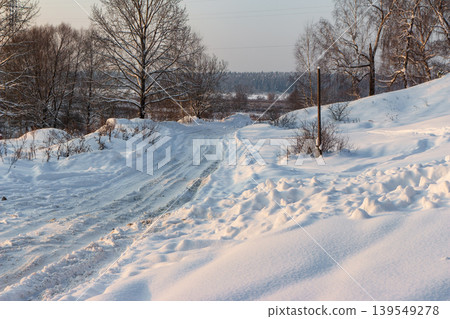 Winter countryside road winding through deep fresh snow at late afternoon golden hour, snowdrifts bathed in warm sunlight, bare trees border the scene Winter countryside road winding through deep fresh snow at late afternoon golden hour, snowdrifts bathed in warm sunlight, bare trees border the scene 139549278