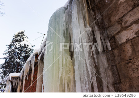 Dramatic, thick icicles cascade down the frosty brick and concrete walls of an old building, capturing winter's stark, frozen beauty. Clear sky and snowy tree 139549296