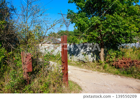 A rustic, weathered gate post stands beside a winding dirt road, flanked by lush green foliage. A tall power line tower looms in the distant background under a bright blue sky A rustic, weathered gate post stands beside a winding dirt road, flanked by lush green foliage. A tall power line tower looms in the distant background under a bright blue sky 139550209