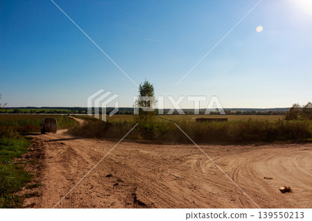 A dusty white van motors down a rural dirt track, stirring up soil as it heads into the scenic countryside. Fields stretch under a bright blue sky, hinting at a journey ahead 139550213