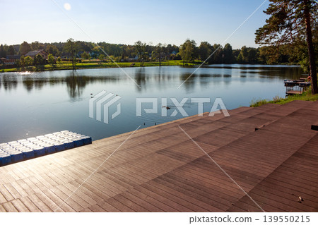 Sunny day view of a wide wooden stepped promenade leading to a placid lake. A blue modular pontoon floats nearby, perfect for waterside relaxation and enjoyment Sunny day view of a wide wooden stepped promenade leading to a placid lake. A blue modular pontoon floats nearby, perfect for waterside relaxation and enjoyment 139550215