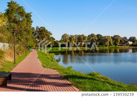 A tranquil park path borders a calm lake under a bright blue sky. Lush greenery lines the walk, inviting a peaceful stroll through nature. Sunbeams dappling the brickway add to the serene atmosphere A tranquil park path borders a calm lake under a bright blue sky. Lush greenery lines the walk, inviting a peaceful stroll through nature. Sunbeams dappling the brickway add to the serene atmosphere 139550239