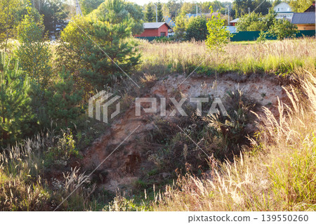 Sunny view of an eroded bank brimming with golden dry grasses, overlooking a peaceful rural village with houses and trees. Serene natural beauty Sunny view of an eroded bank brimming with golden dry grasses, overlooking a peaceful rural village with houses and trees. Serene natural beauty 139550260
