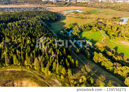 Aerial view of a sprawling green forest bordering a meandering river, with fields and distant urban sprawl under warm sunlight. A serene natural landscape in late summer Aerial view of a sprawling green forest bordering a meandering river, with fields and distant urban sprawl under warm sunlight. A serene natural landscape in late summer 139550273